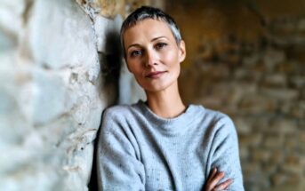 A person with short gray hair and a gray sweater stands with arms crossed, leaning against a stone wall, looking calmly at the camera.