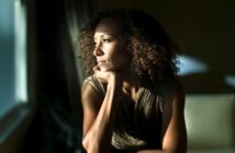 A woman with curly hair rests her chin on her hand, gazing thoughtfully out a window with sunlight illuminating her face.