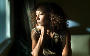 A woman with curly hair rests her chin on her hand, gazing thoughtfully out a window with sunlight illuminating her face.