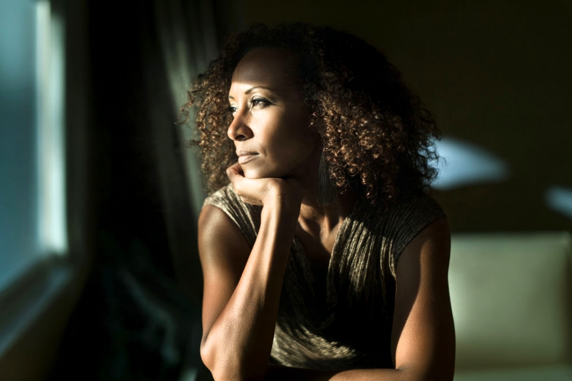 A woman with curly hair rests her chin on her hand, gazing thoughtfully out a window with sunlight illuminating her face.