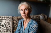 A woman with light hair and blue eyes sits on a patterned sofa, wearing a blue patterned blouse, looking calmly at the camera in a softly lit room.