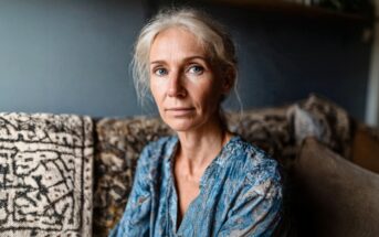 A woman with light hair and blue eyes sits on a patterned sofa, wearing a blue patterned blouse, looking calmly at the camera in a softly lit room.