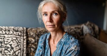 A woman with light hair and blue eyes sits on a patterned sofa, wearing a blue patterned blouse, looking calmly at the camera in a softly lit room.