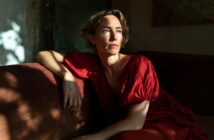 A woman in a red dress sits on a sofa, looking thoughtfully into the sunlight streaming through a window, casting shadows on the wall and her face.