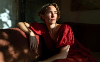 A woman in a red dress sits on a sofa, looking thoughtfully into the sunlight streaming through a window, casting shadows on the wall and her face.