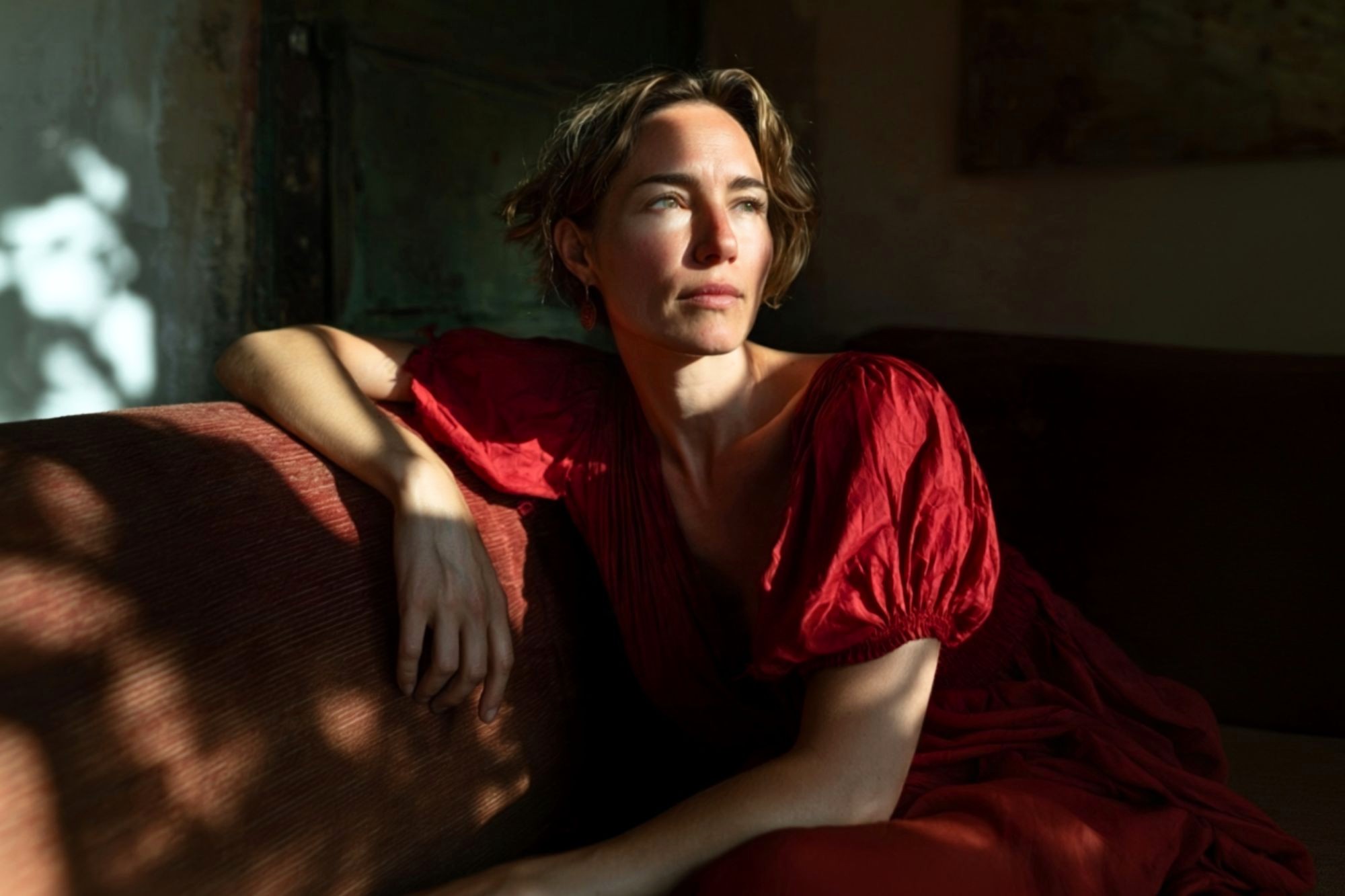 A woman in a red dress sits on a sofa, looking thoughtfully into the sunlight streaming through a window, casting shadows on the wall and her face.