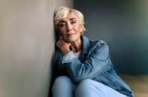 Older woman with short blonde hair and a denim jacket sits on the floor, leaning against a wall and looking thoughtfully at the camera in soft, natural light.