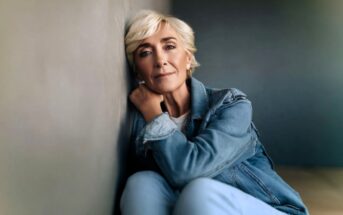 Older woman with short blonde hair and a denim jacket sits on the floor, leaning against a wall and looking thoughtfully at the camera in soft, natural light.