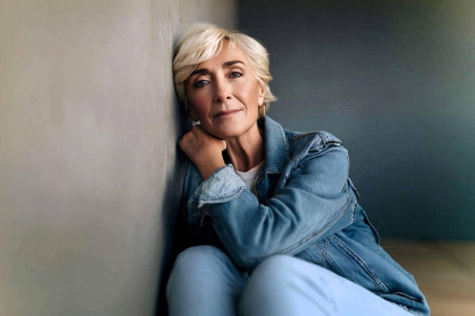 Older woman with short blonde hair and a denim jacket sits on the floor, leaning against a wall and looking thoughtfully at the camera in soft, natural light.