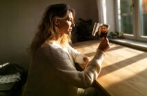 A woman in a white sweater sits at a wooden table by a window, holding and looking thoughtfully at a small photo of two people smiling together. Sunlight streams in, creating a warm, reflective atmosphere.