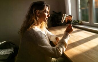 A woman in a white sweater sits at a wooden table by a window, holding and looking thoughtfully at a small photo of two people smiling together. Sunlight streams in, creating a warm, reflective atmosphere.