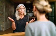 A woman with gray hair looks concerned and gestures with her hands while talking to another woman, who is seen from behind. They are indoors, sitting at a table.