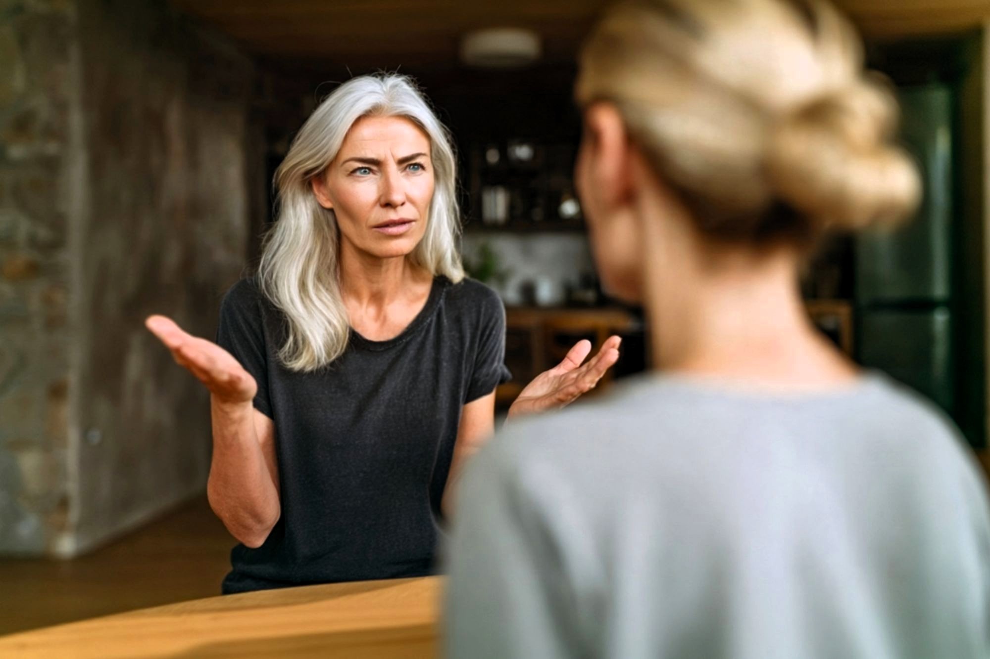 A woman with gray hair looks concerned and gestures with her hands while talking to another woman, who is seen from behind. They are indoors, sitting at a table.