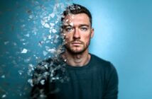 A man with short hair and a beard stares ahead against a blue background, with the left side of his face and body dissolving into shattered glass-like fragments, giving a surreal, disappearing effect.