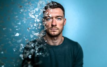 A man with short hair and a beard stares ahead against a blue background, with the left side of his face and body dissolving into shattered glass-like fragments, giving a surreal, disappearing effect.