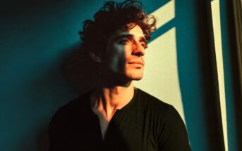 A man with curly hair looks thoughtfully out a window as warm sunlight casts dramatic shadows across his face and a teal wall behind him.