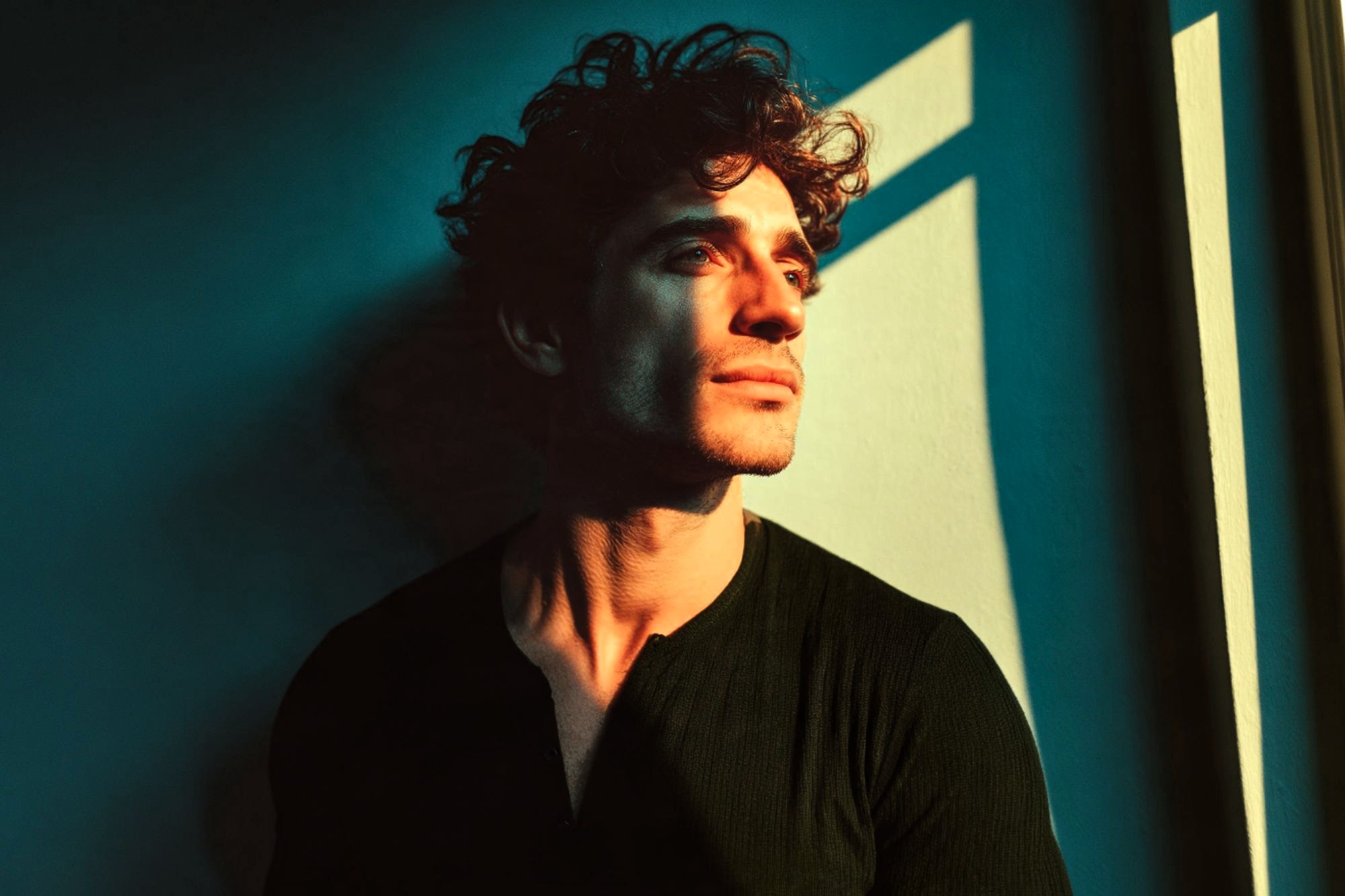 A man with curly hair looks thoughtfully out a window as warm sunlight casts dramatic shadows across his face and a teal wall behind him.