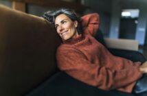 A woman with gray hair in a cozy orange sweater relaxes on a dark sofa, smiling while looking off to the side in a softly lit room.