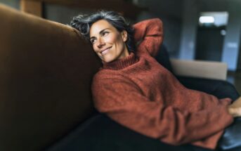 A woman with gray hair in a cozy orange sweater relaxes on a dark sofa, smiling while looking off to the side in a softly lit room.