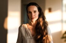 A woman with long wavy brown hair and a white blouse stands indoors, softly lit by natural light, with a calm expression and a blurred background.