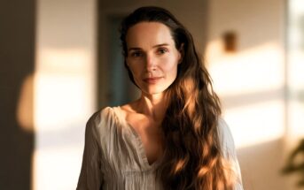A woman with long wavy brown hair and a white blouse stands indoors, softly lit by natural light, with a calm expression and a blurred background.