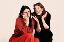 Two women sit together. One looks bored and rests her chin on her hand, wearing red. The other, in black, gestures energetically and smiles, appearing excited or enthusiastic. The background is plain and neutral.