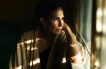 A woman with dark hair pulled back gazes thoughtfully out a window, resting her head on her hand. Soft sunlight and shadows fall across her face and beige shawl, creating a contemplative mood.