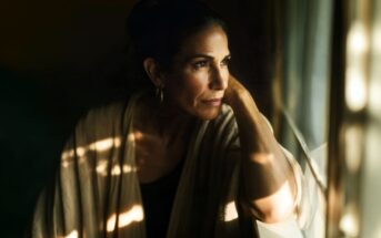 A woman with dark hair pulled back gazes thoughtfully out a window, resting her head on her hand. Soft sunlight and shadows fall across her face and beige shawl, creating a contemplative mood.