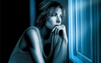 A woman with short hair gazes thoughtfully out a window at night, resting her chin on her hand. Blue lighting creates a contemplative mood.