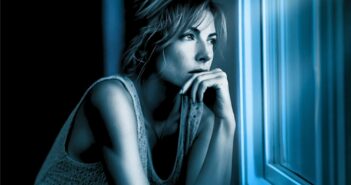 A woman with short hair gazes thoughtfully out a window at night, resting her chin on her hand. Blue lighting creates a contemplative mood.