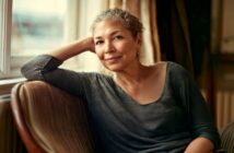A woman with short curly gray hair sits on a striped chair by a window, smiling softly, with her elbow resting on the chair’s back and her head leaning on her hand. She wears a loose dark gray top.