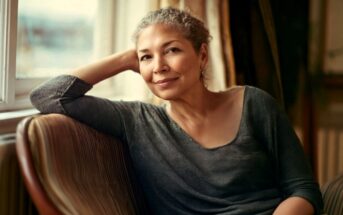 A woman with short curly gray hair sits on a striped chair by a window, smiling softly, with her elbow resting on the chair’s back and her head leaning on her hand. She wears a loose dark gray top.