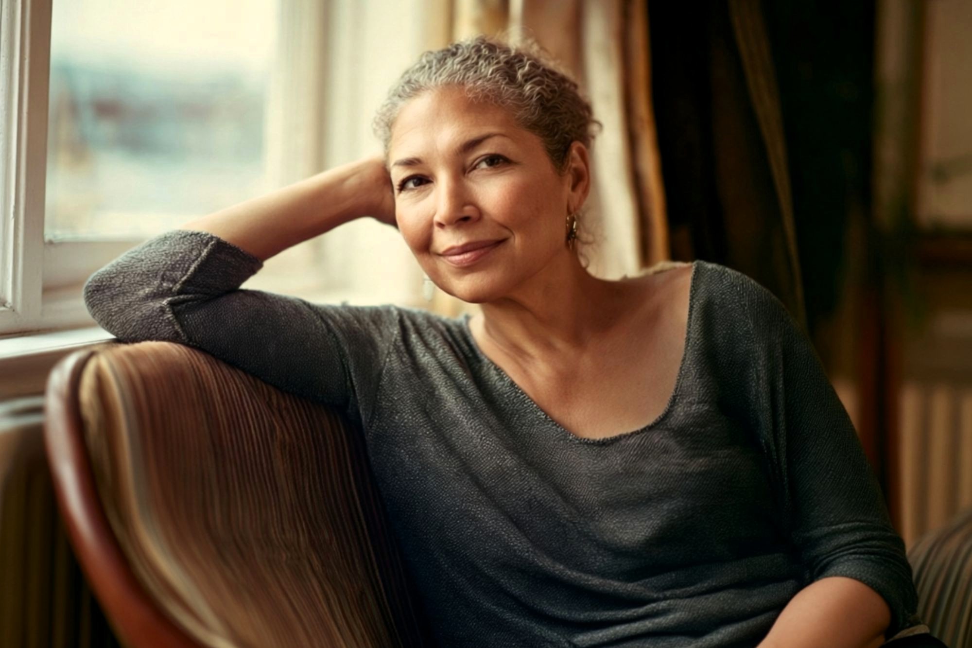 A woman with short curly gray hair sits on a striped chair by a window, smiling softly, with her elbow resting on the chair’s back and her head leaning on her hand. She wears a loose dark gray top.