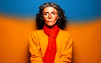 A woman with curly gray hair and glasses wears an orange coat and red scarf, standing against a blue and orange background, looking directly at the camera.