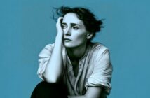 A person with short, tousled hair sits against a blue background, resting their head on one hand and looking thoughtful or pensive, wearing a loose, buttoned shirt.