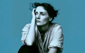 A person with short, tousled hair sits against a blue background, resting their head on one hand and looking thoughtful or pensive, wearing a loose, buttoned shirt.