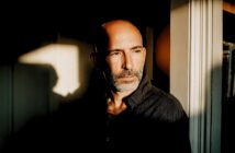 A middle-aged man with a shaved head and beard stands by a sunlit window, looking thoughtfully into the distance. Shadows and warm light fall across his face and dark shirt.