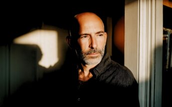 A middle-aged man with a shaved head and beard stands by a sunlit window, looking thoughtfully into the distance. Shadows and warm light fall across his face and dark shirt.