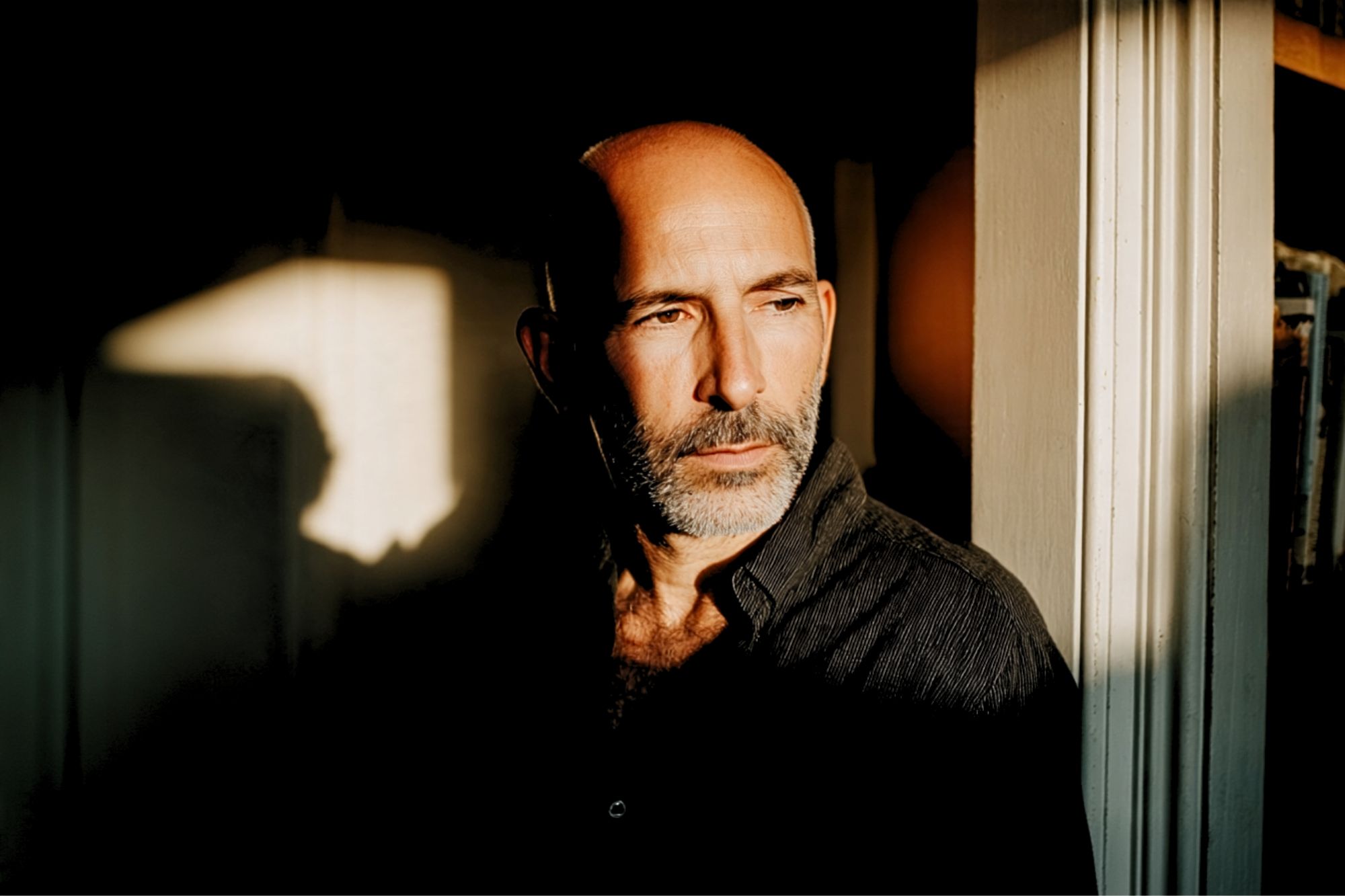 A middle-aged man with a shaved head and beard stands by a sunlit window, looking thoughtfully into the distance. Shadows and warm light fall across his face and dark shirt.