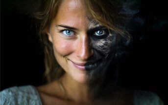 A woman with one side of her face smiling in natural light and the other side darkened, appearing cracked and smoky, creating a contrast between vitality and decay against a black background.