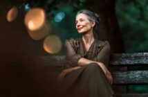 A woman with gray hair and red lipstick sits on a wooden bench outdoors, smiling softly and looking to the side, with blurred lights and greenery in the background.