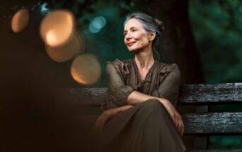 A woman with gray hair and red lipstick sits on a wooden bench outdoors, smiling softly and looking to the side, with blurred lights and greenery in the background.