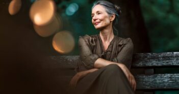 A woman with gray hair and red lipstick sits on a wooden bench outdoors, smiling softly and looking to the side, with blurred lights and greenery in the background.
