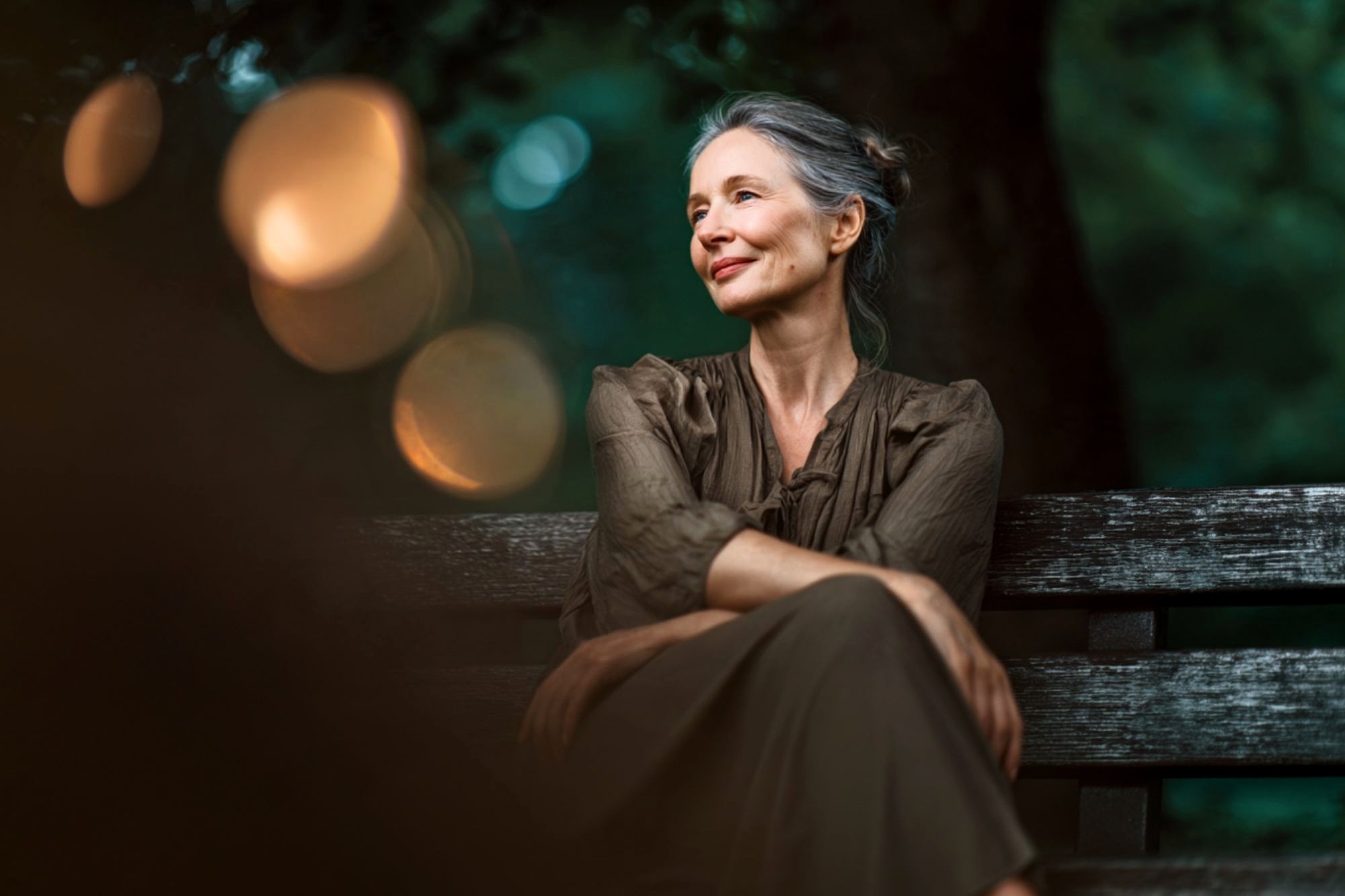 A woman with gray hair and red lipstick sits on a wooden bench outdoors, smiling softly and looking to the side, with blurred lights and greenery in the background.