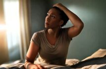 A woman sits on a bed in a softly lit room, looking up with a worried expression while holding her head with one hand. Sunlight streams through a window, casting a gentle glow on her face.