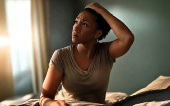 A woman sits on a bed in a softly lit room, looking up with a worried expression while holding her head with one hand. Sunlight streams through a window, casting a gentle glow on her face.