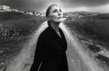 An older woman in a dark outfit stands at a rural crossroads, gazing upward. The photo is black and white, with distant buildings and cloudy skies in the background.