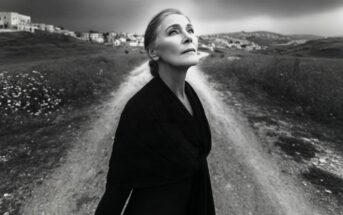 An older woman in a dark outfit stands at a rural crossroads, gazing upward. The photo is black and white, with distant buildings and cloudy skies in the background.