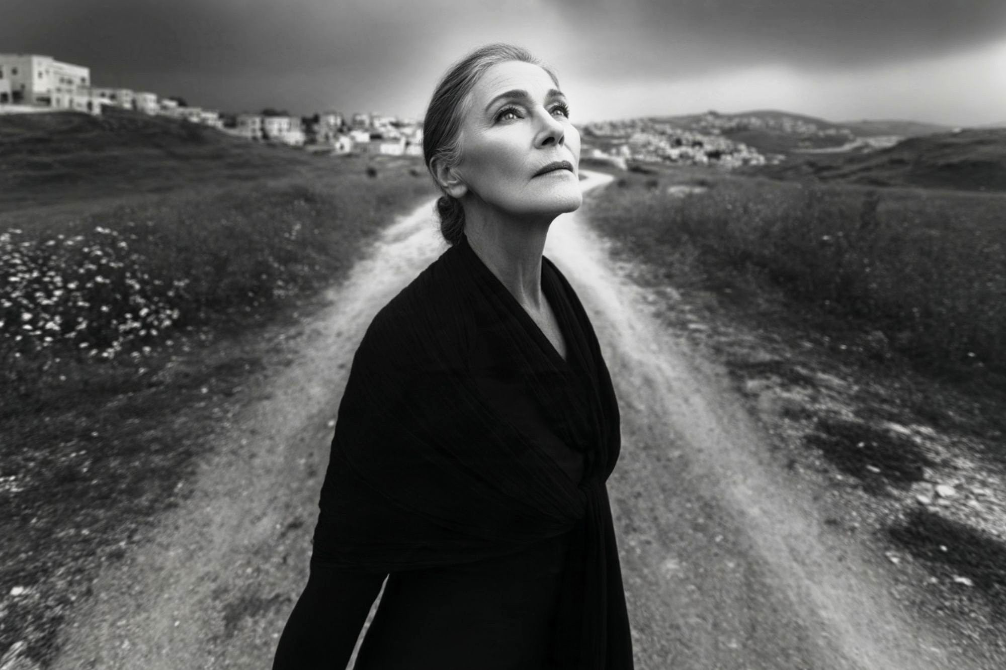 An older woman in a dark outfit stands at a rural crossroads, gazing upward. The photo is black and white, with distant buildings and cloudy skies in the background.