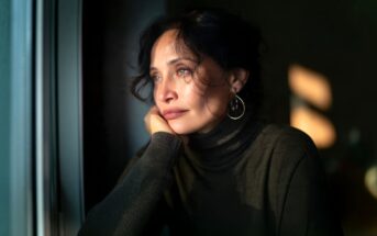 Woman with dark hair and hoop earrings gazes thoughtfully out a window, resting her chin on her hand. She wears a dark turtleneck, and soft sunlight falls across her face. The mood appears pensive and reflective.